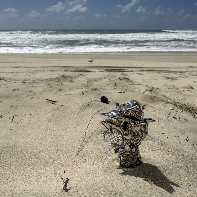 The silver pendant sample standing in the sand, with the open sea breaking behind it
